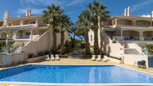 Zona de piscina con una piscina azul cristalina, sillones blancos, palmeras y una escalera que conduce a un edificio con balcones.