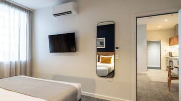 A hotel room with a bed, a TV, a mirror, and a kitchenette in the background.