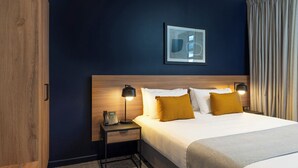 Una habitación de hotel con cabecera de madera, una mesita de noche con lámpara y una cama con almohadas blancas y mostaza.