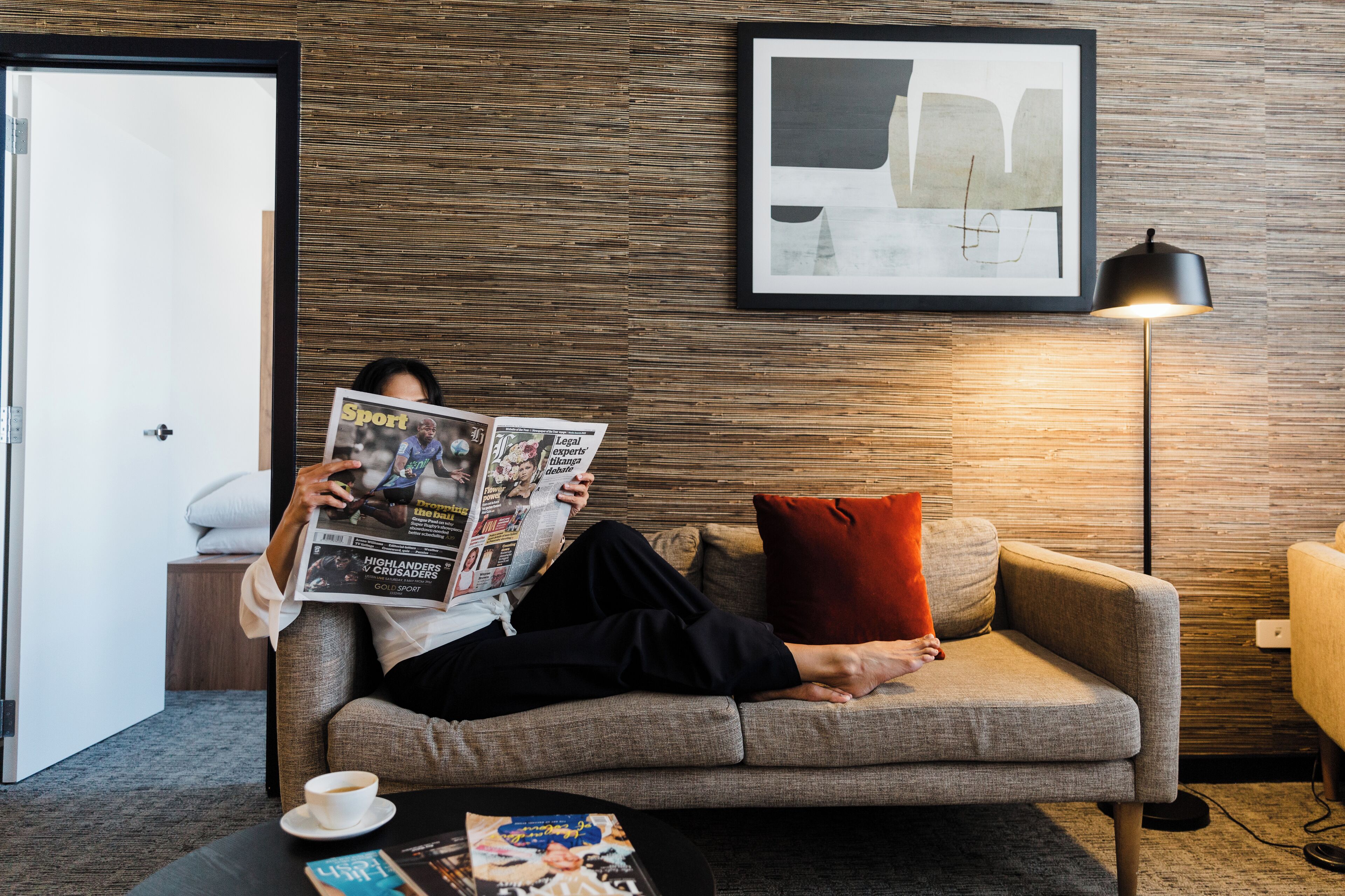 Una persona leyendo una revista en un sofá en una habitación con pared de madera, una obra de arte abstracta enmarcada y una lámpara de pie.