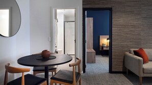 Una habitación de hotel moderna con una mesa de comedor redonda, dos sillas de madera, un sofá con un cojín rojo, un espejo grande y vistas de la ciudad al atardecer.