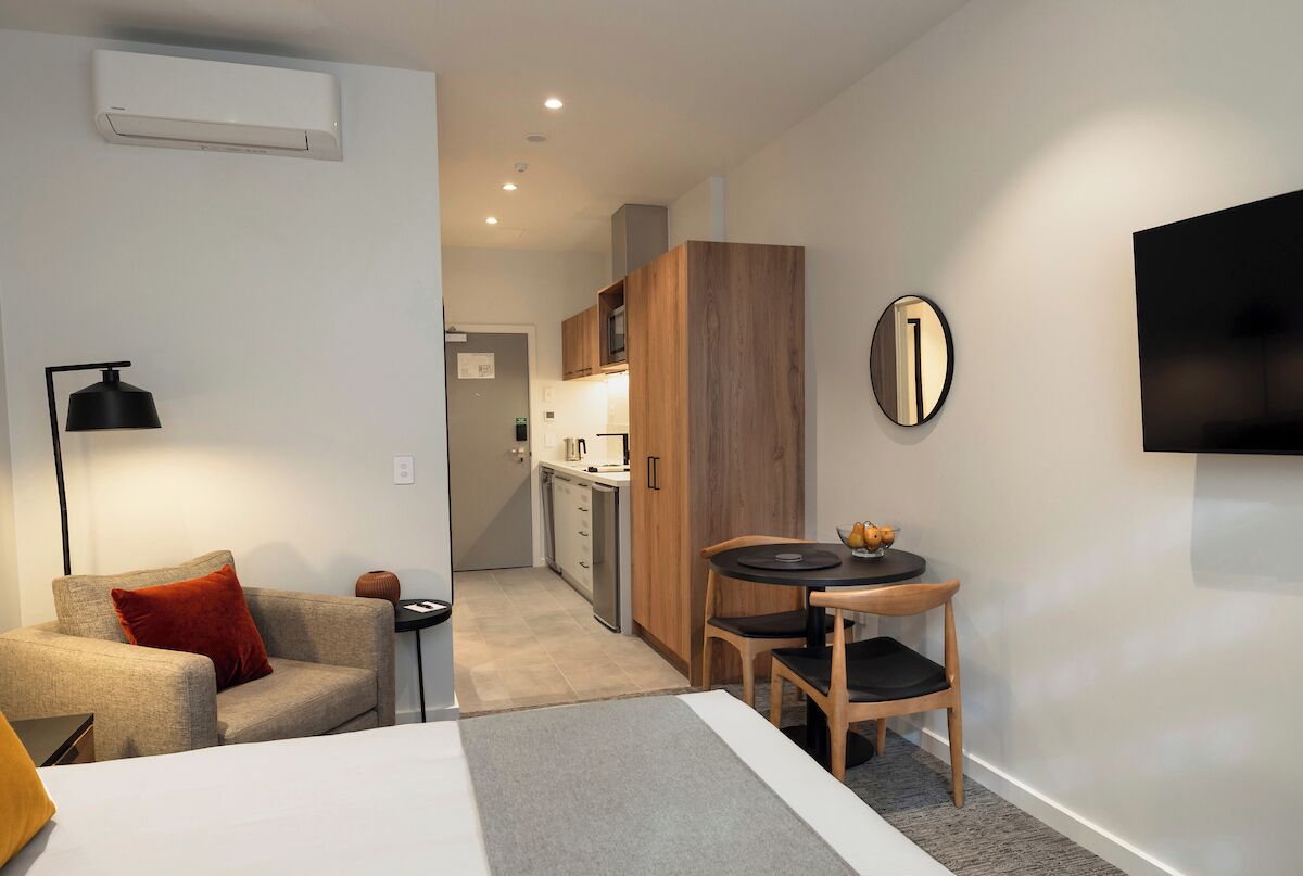 Een moderne hotelkamer met een bed, een klein bureau, een stoel, een televisie en een kitchenette op de achtergrond.