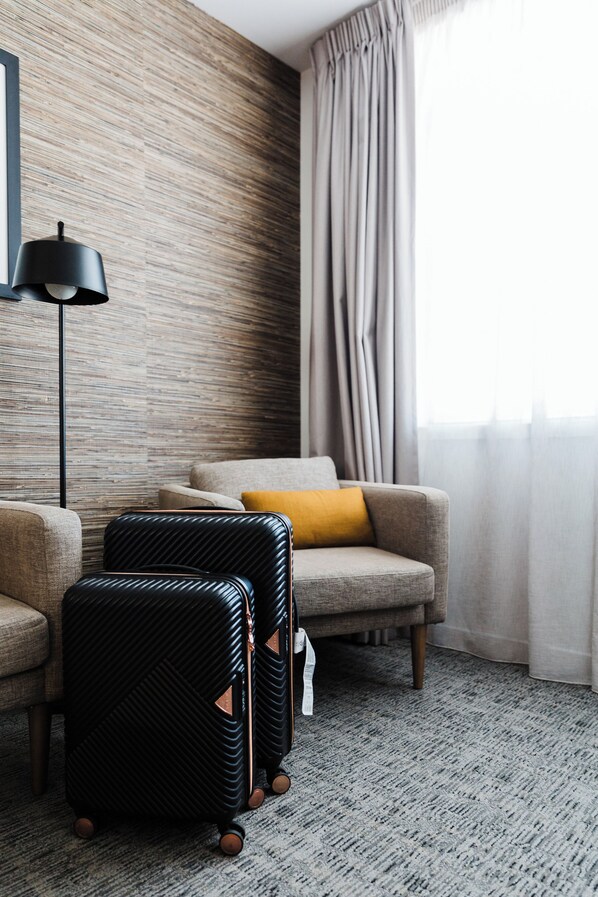 Habitación de hotel con sofá gris, maleta negra, lámpara y ventana con cortinas.