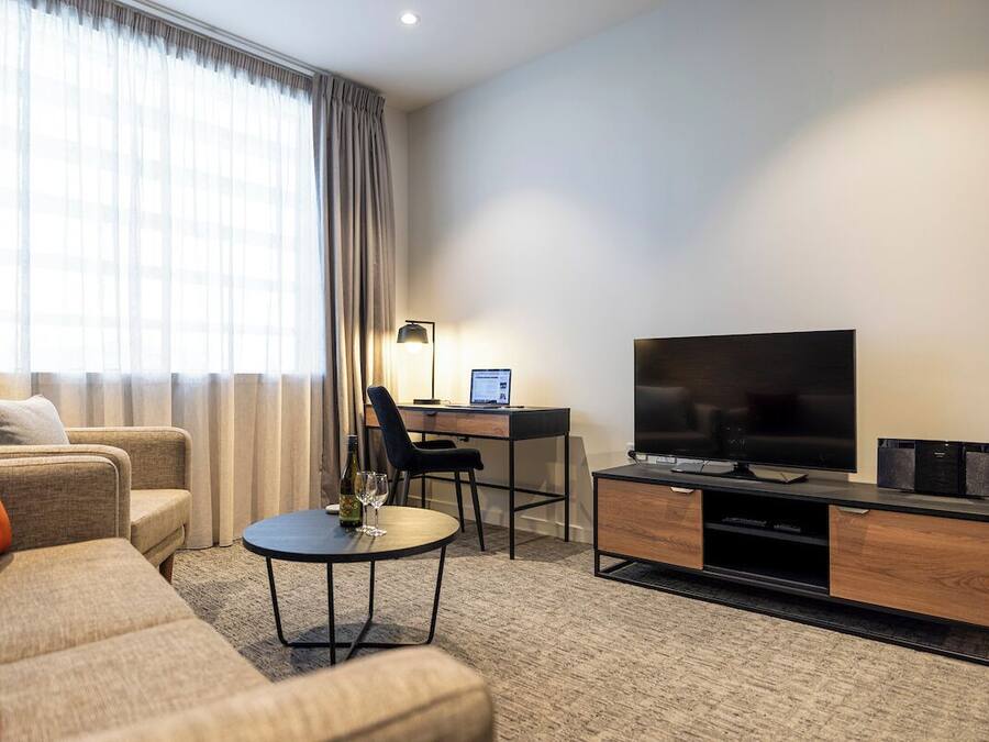 Una habitación de hotel moderna con sofá, escritorio, ordenador portátil, televisor y mesa de centro.