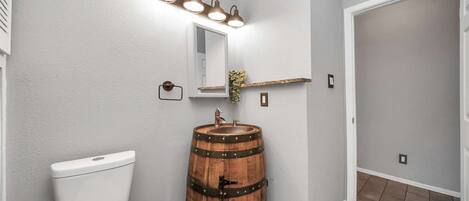 Un baño con inodoro, lavamanos de barril de madera, espejo y lámpara sobre el espejo.