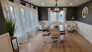 A dining area with a wooden table and chairs, a chandelier, and a large window with curtains. - Stunning and Completely Remodeled Ranch Home! Walk to Downtown & Marina! (Port Washington)