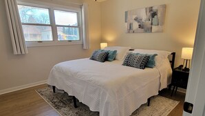 A bedroom with a large bed, two bedside lamps, a window with curtains, and a painting on the wall. - Stunning and Completely Remodeled Ranch Home! Walk to Downtown & Marina! (Port Washington)