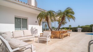 A sunlit patio with a white sofa, wooden tables, and chairs, palm trees, and a swimming pool. - Gorgeous home in Marbella (Marbella)