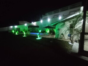 A nighttime view of a hotel with a pool area, outdoor seating, and illuminated plants.