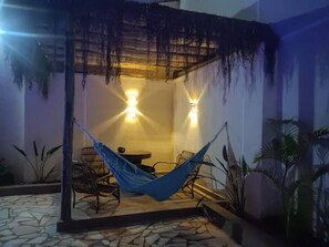 A hammock under a pergola with two wall-mounted lights, a small table, and chairs in a courtyard with tiled flooring and potted plants.