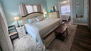A bedroom with a bed, bedside tables, a dresser, a bench, a mirror, and a door leading to another room. - Aunt May's - Waterfront Cottage in Cedar Key! (Cedar Key)