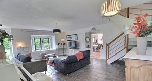 A spacious living room with a piano, a grey sofa, a television, and a staircase leading to another level.