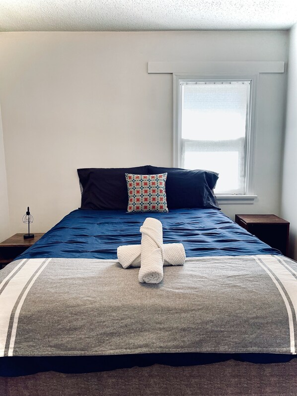 A neatly made bed with a blue bedspread, two pillows, and a rolled towel. - 2-Bedroom Downtown Suite | King Bed (Weyburn)