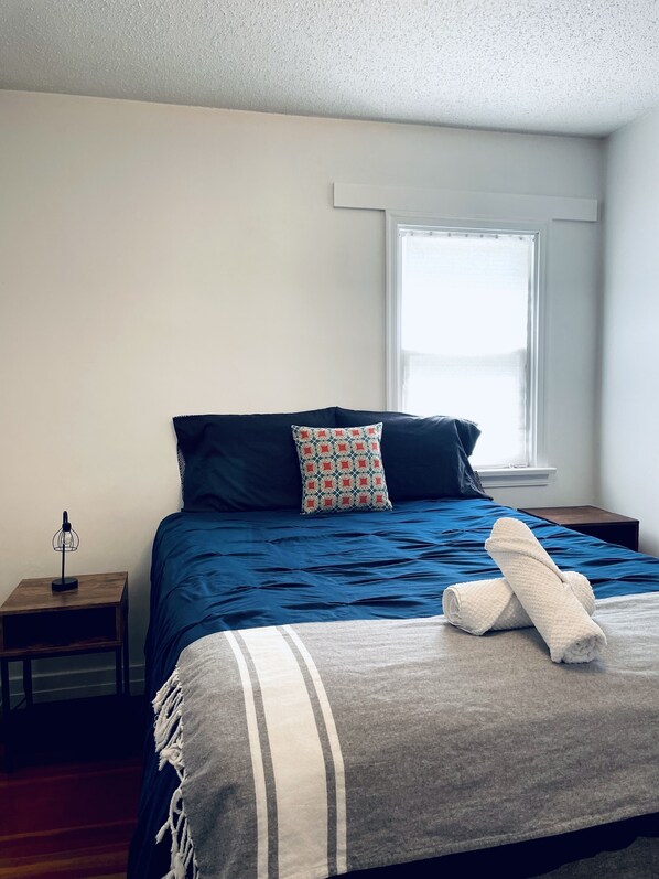 Una cama bien hecha con una colcha azul y un cojín estampado, una mesita de noche con una lámpara y una ventana con persianas.