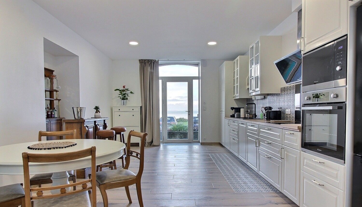 A modern kitchen with white cabinets, a dining area with wooden chairs, and a large window offering a view of the outdoors.