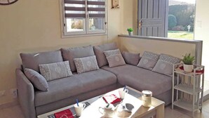 A living room with a grey sofa, a coffee table, a clock, and a window with blinds. - Gîtes de France® - Les Hirondelles (ST REMY BOSCROCOURT)