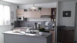 A modern kitchen with a central island, wooden cabinets, a microwave, and a hanging pendant light. - Gîtes de France® - Les Hirondelles (ST REMY BOSCROCOURT)