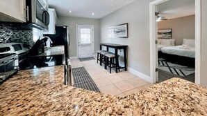 A modern kitchen with granite countertops, a dining area with a table and chairs, a small seating area with a bench, and a bedroom with a bed and a nightstand.
