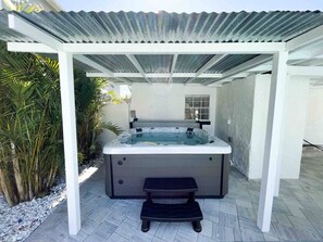 Un jacuzzi bajo una pérgola de paredes blancas, rodeado de plantas y un patio con baldosas.