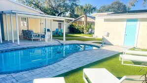 A swimming pool with a covered patio area, a bathroom, and a lawn with lounge chairs.