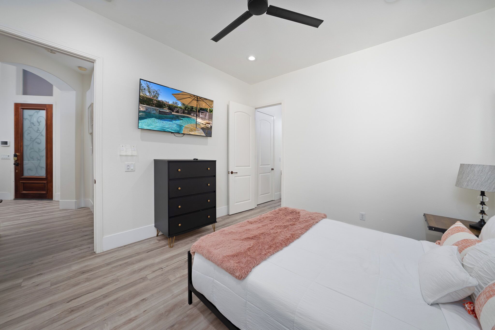 Une chambre à coucher comprenant un lit, un ventilateur de plafond, un téléviseur fixé au mur, une commode et une porte.