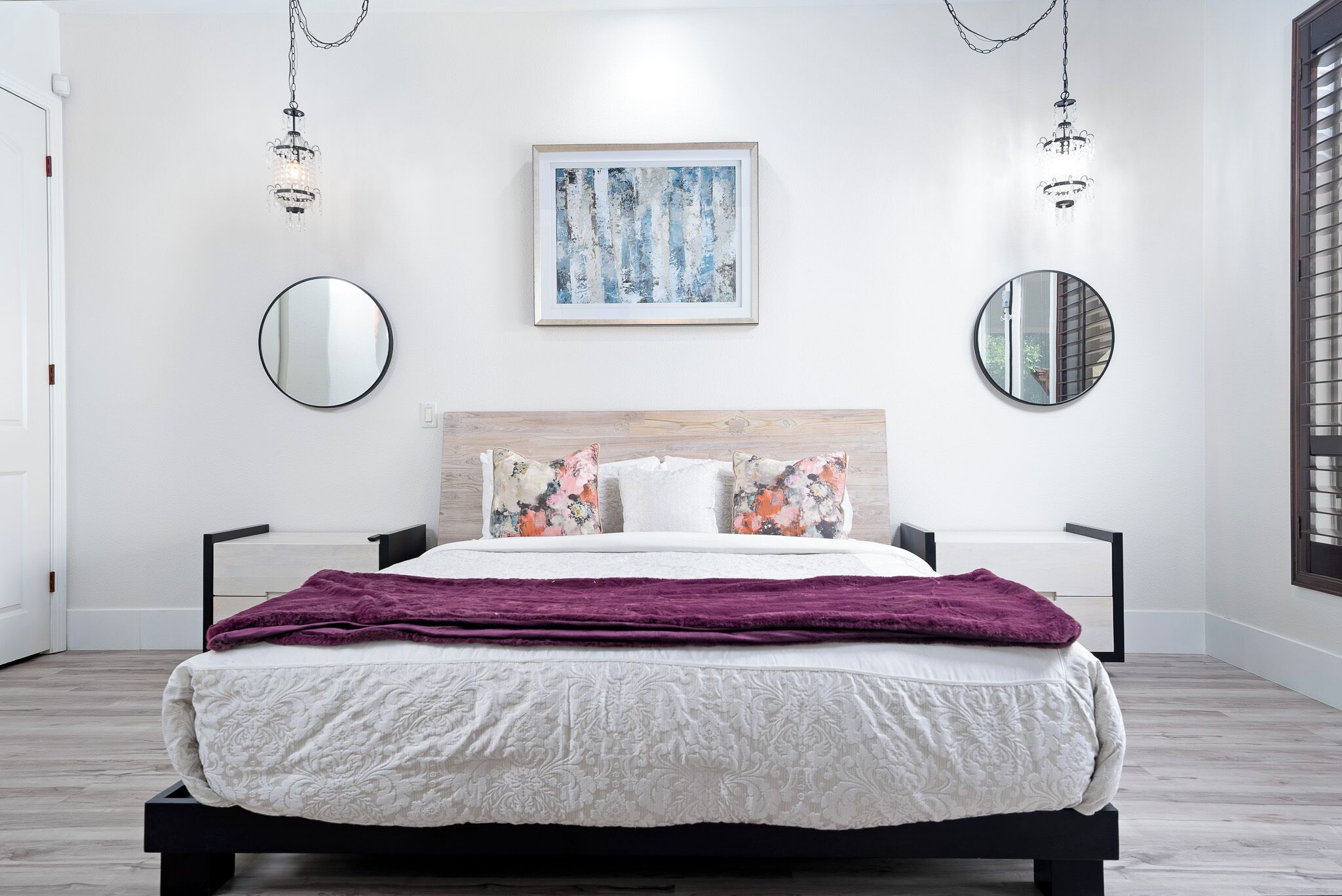 A modern bedroom with a bed, two wall-mounted mirrors, a framed abstract painting, and a chandelier.