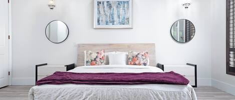 A modern bedroom with a bed, two wall-mounted mirrors, a framed abstract painting, and a chandelier.