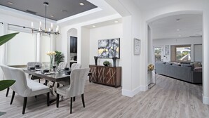 A modern dining area with a wooden table, white upholstered chairs, and a chandelier. - Casa Serenity Luxury Living w/ Pool & Jacuzzi (Indio)