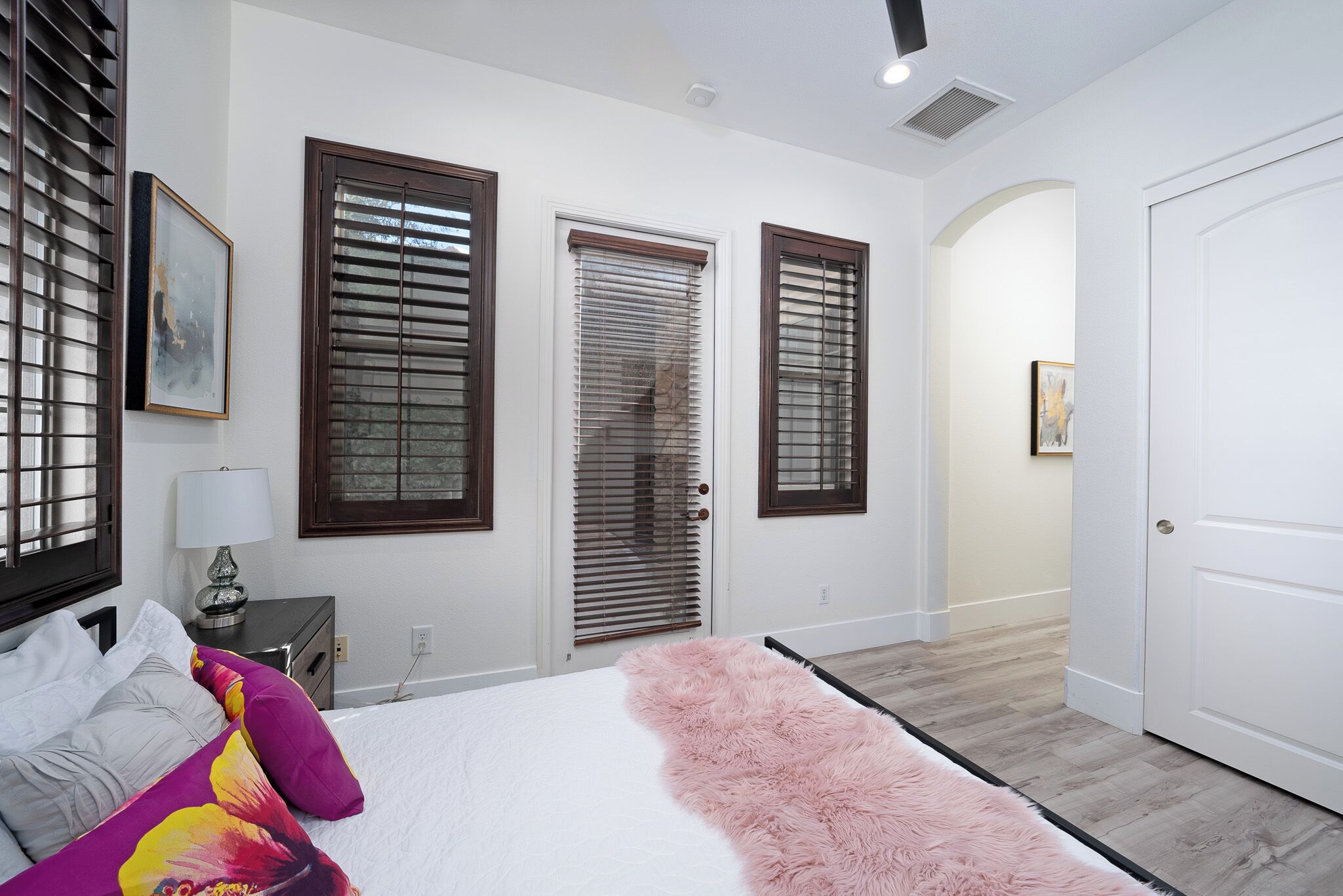 A bedroom with a bed, a fluffy pink rug, and three windows with blinds.