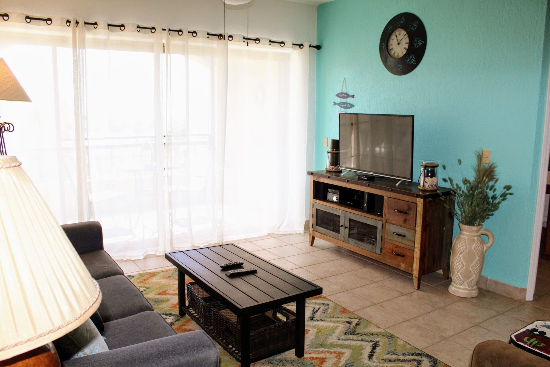A living room with a sofa, a coffee table, a TV on a wooden stand, and a large window with curtains.