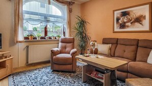 En stue med en brun lædersofa, en brun lænestol, et træbord og et stort vindue med gardiner. Der står plantekasser på vindueskarmen, og der hænger et billede i en ramme på væggen.