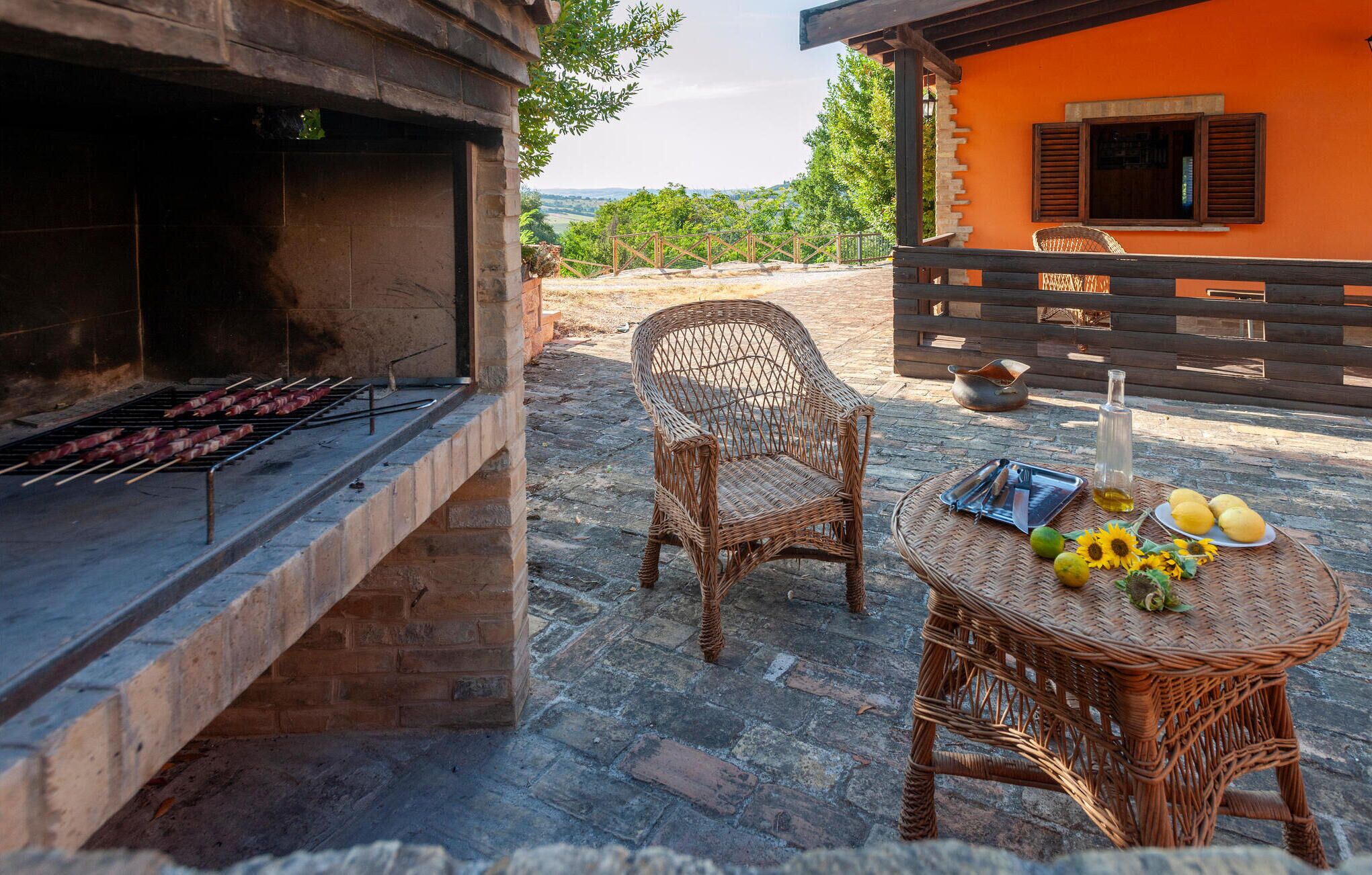 An outdoor barbecue area with a grill, wicker chairs, and a table with food and drinks.