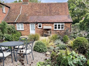 Een stenen huis met pannendak, een tuin met diverse planten, een barbecueplek en een tafel met stoelen.