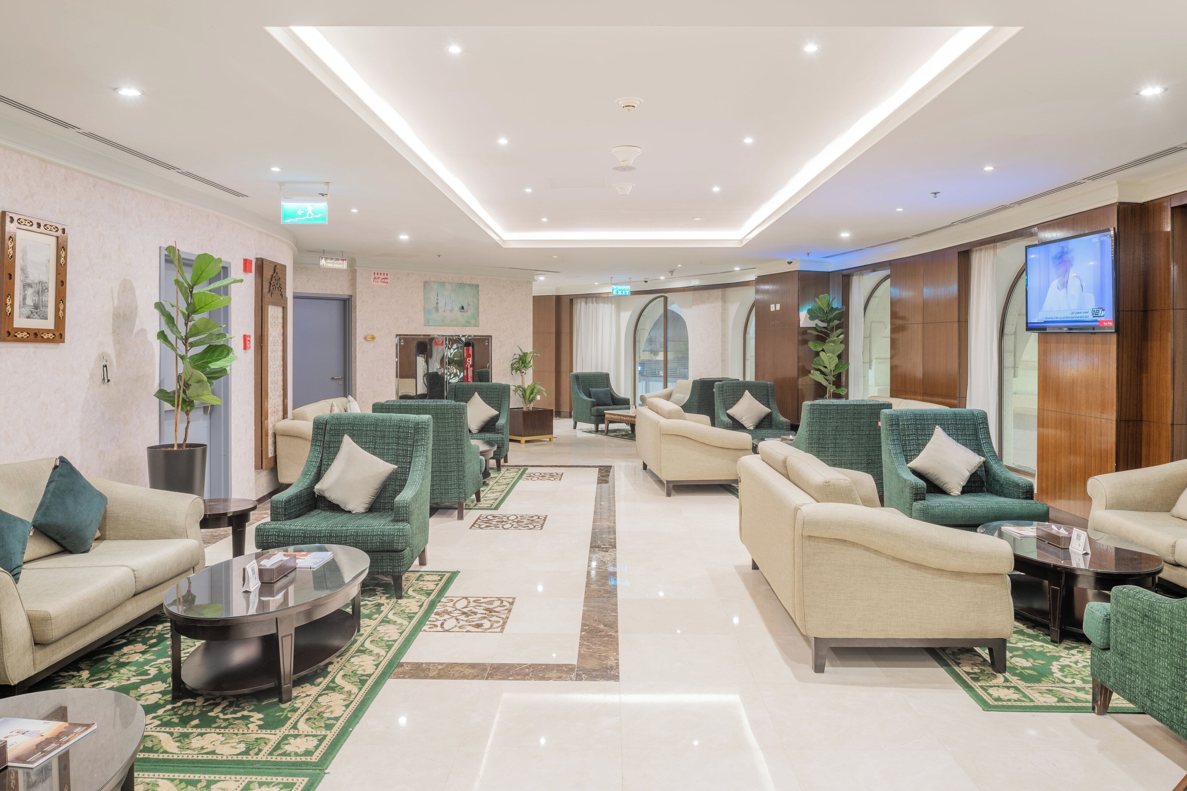Un moderno vestíbulo de hotel con una mezcla de sofás verde y beige, un televisor de pantalla plana y una zona de recepción con un escritorio de madera.