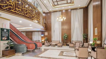 Un vestíbulo de hotel con una zona de estar central, una escalera con ascensor mecánico y un candelabro.