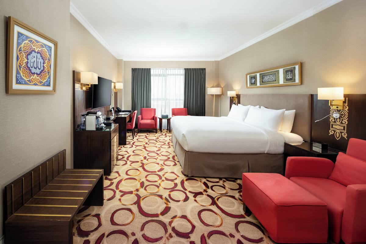 Une chambre d’hôtel avec un lit, un bureau avec un ordinateur, une télévision, un canapé rouge et un tapis orné d’un motif circulaire.