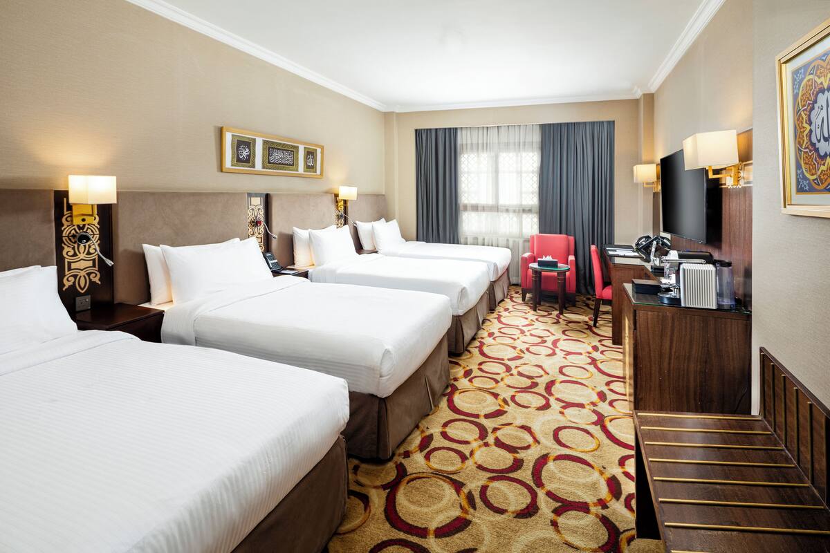Une chambre d’hôtel avec trois lits, un fauteuil rouge, un bureau avec un ordinateur et une télévision.
