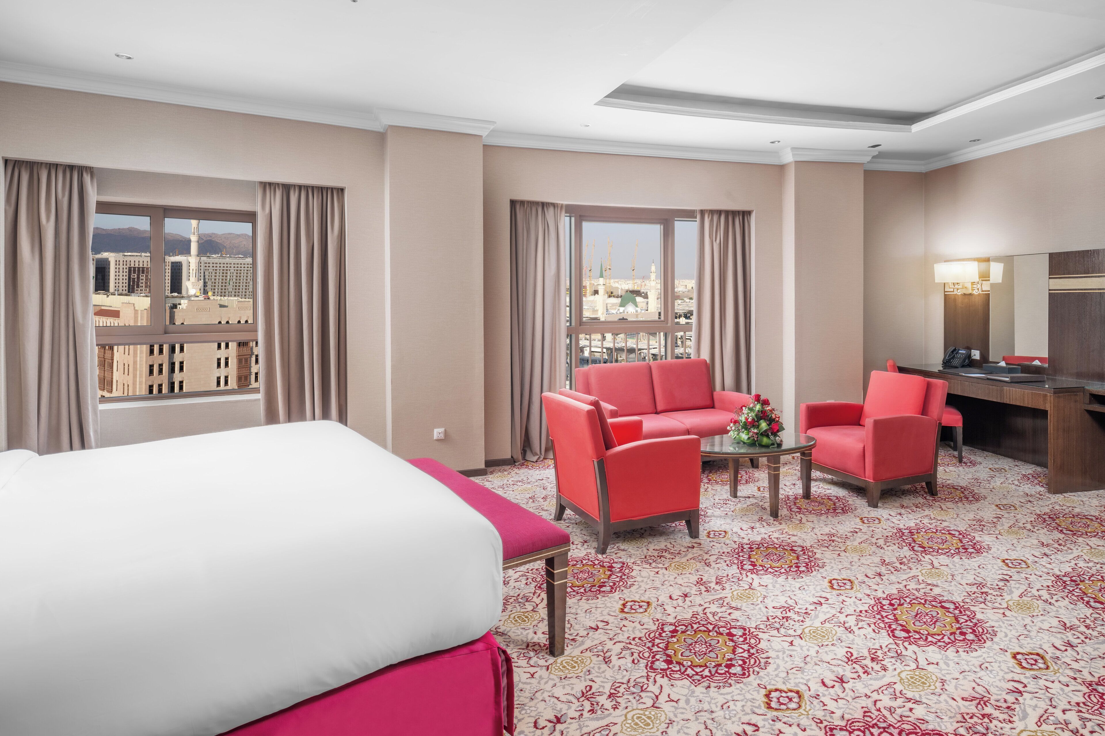 A hotel room with a bed, two red armchairs, a small table with a flower, and a desk with a lamp.