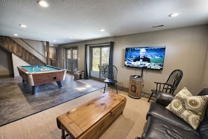 A living room with a pool table, a wooden coffee table, a leather couch, and a television displaying a sports event.