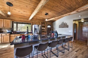 A kitchen with wooden beams, a stainless steel refrigerator, a large island with bar stools, and a window overlooking trees.
