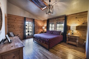 Une chambre avec un plancher en bois, un lit recouvert d’une couverture à carreaux, une table de chevet avec une lampe et une fenêtre avec des rideaux.