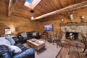 A log cabin interior with a stone fireplace, leather furniture, and a mounted deer head.