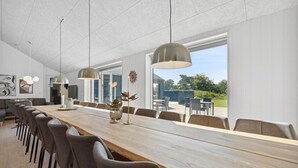 A modern dining area with a long wooden table, multiple chairs, pendant lighting, and large windows offering a view of the outdoors. - Pet friendly home in Rømø with sauna (Rømø)