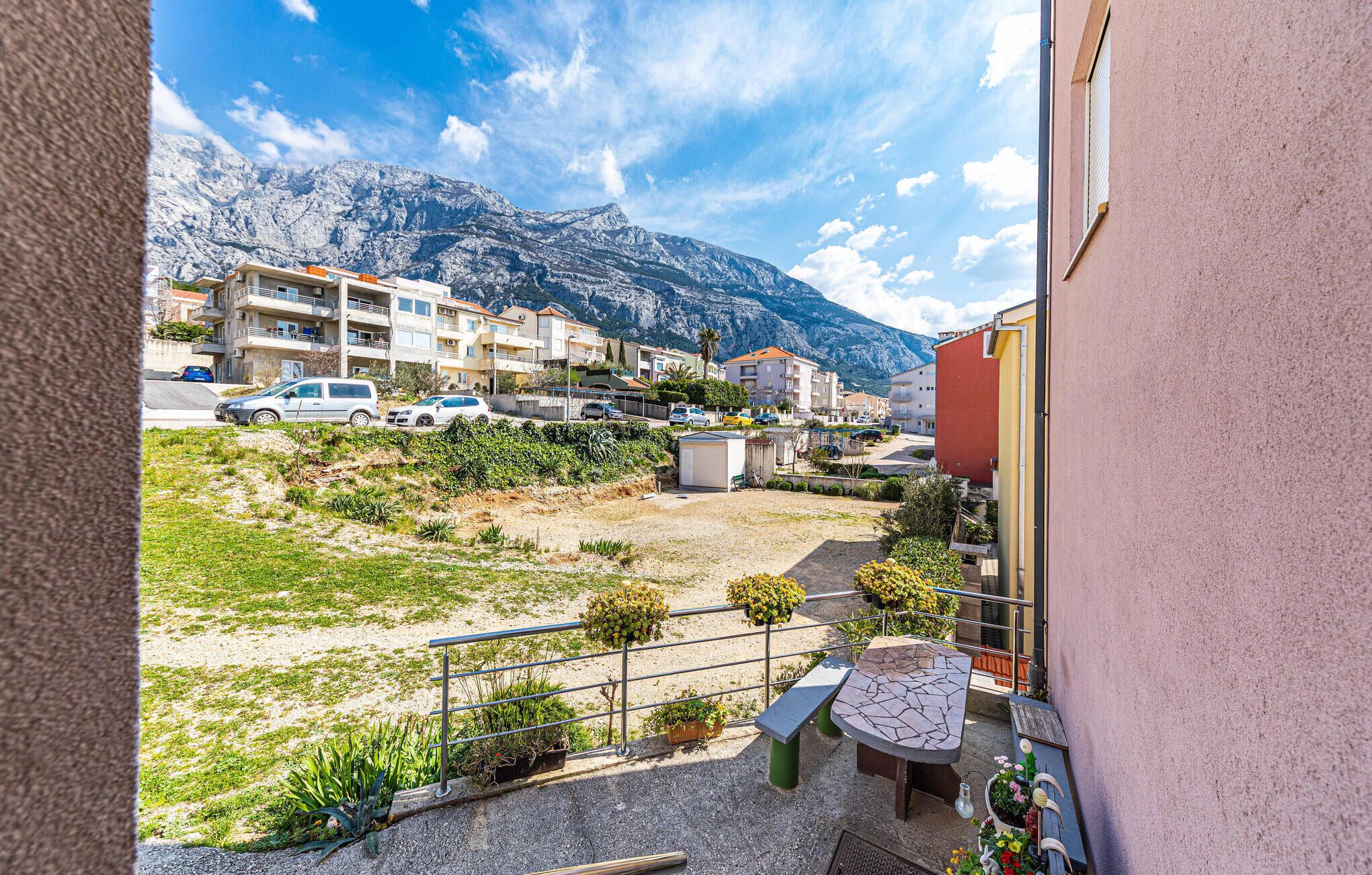 Un balcon avec une table et des chaises, donnant sur un parking et sur des bâtiments, avec une chaîne de montagnes en arrière-plan.