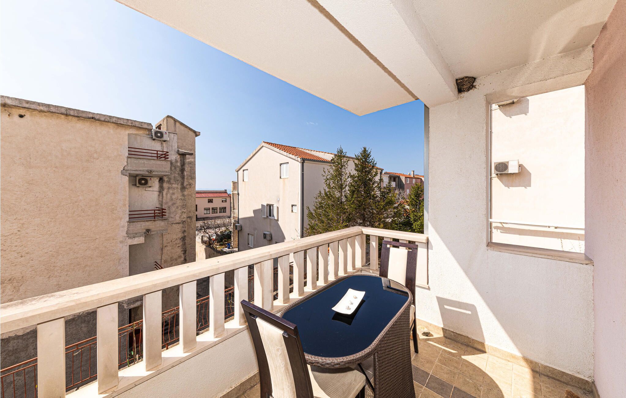 A balcony with a table and chair, overlooking a residential area with buildings and a tree.