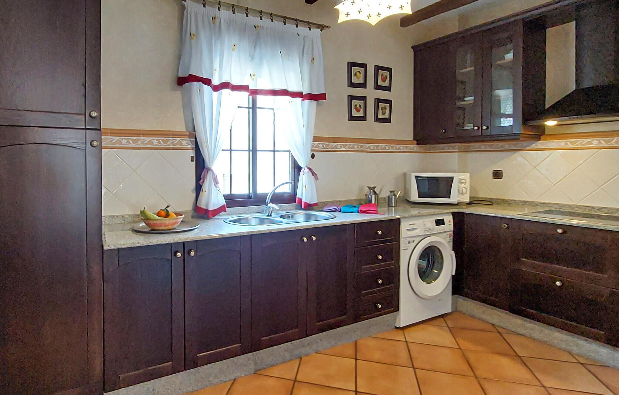 A kitchen with dark wooden cabinets, a washing machine, a microwave, and a window with curtains.