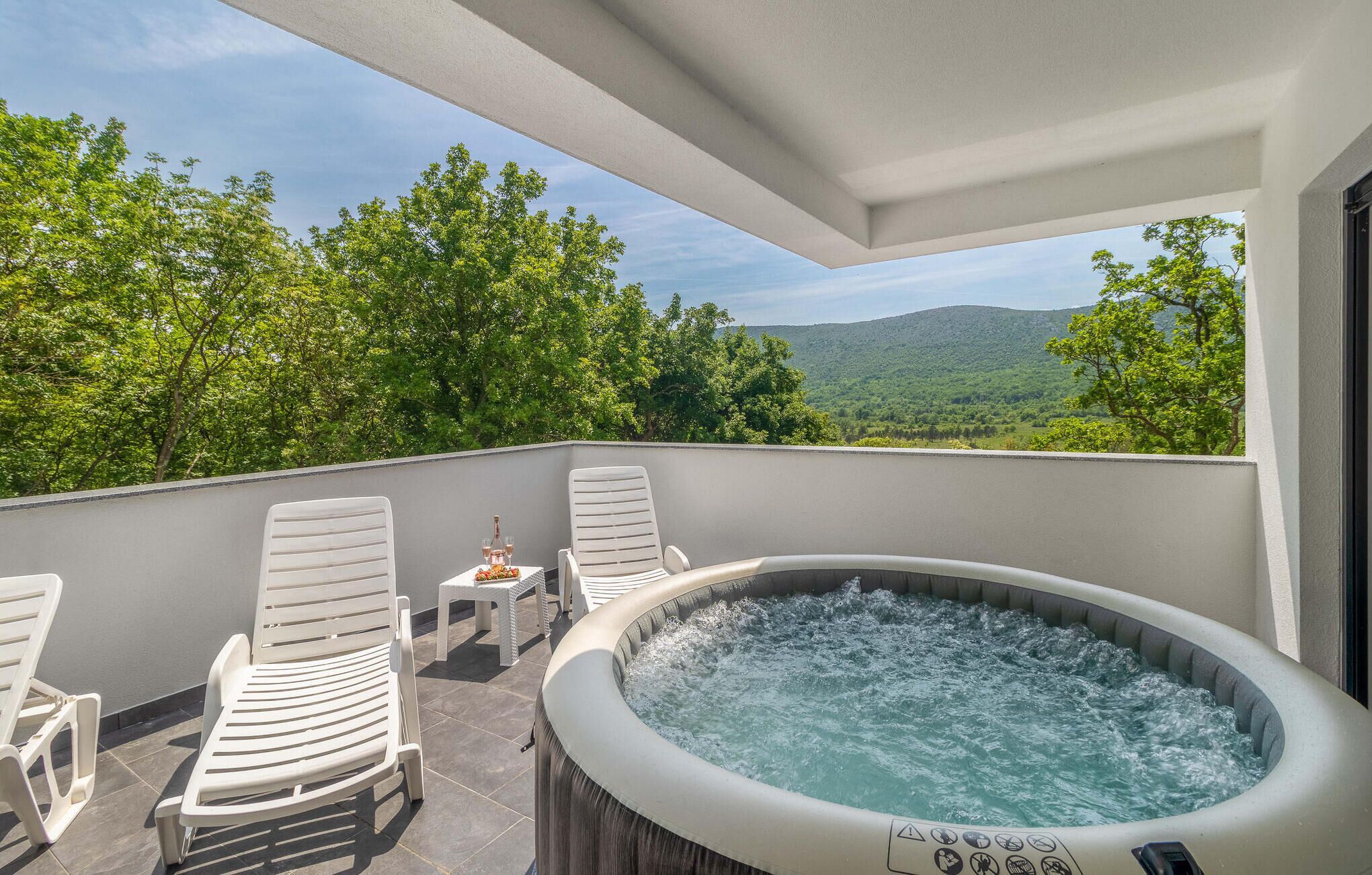Un jacuzzi sur un balcon, avec des chaises et une table blanches, entouré d’arbres et offrant une vue sur les montagnes.