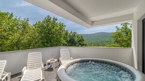 Un jacuzzi en un balcón con sillas blancas y una mesa, rodeado de árboles y vistas a montañas.