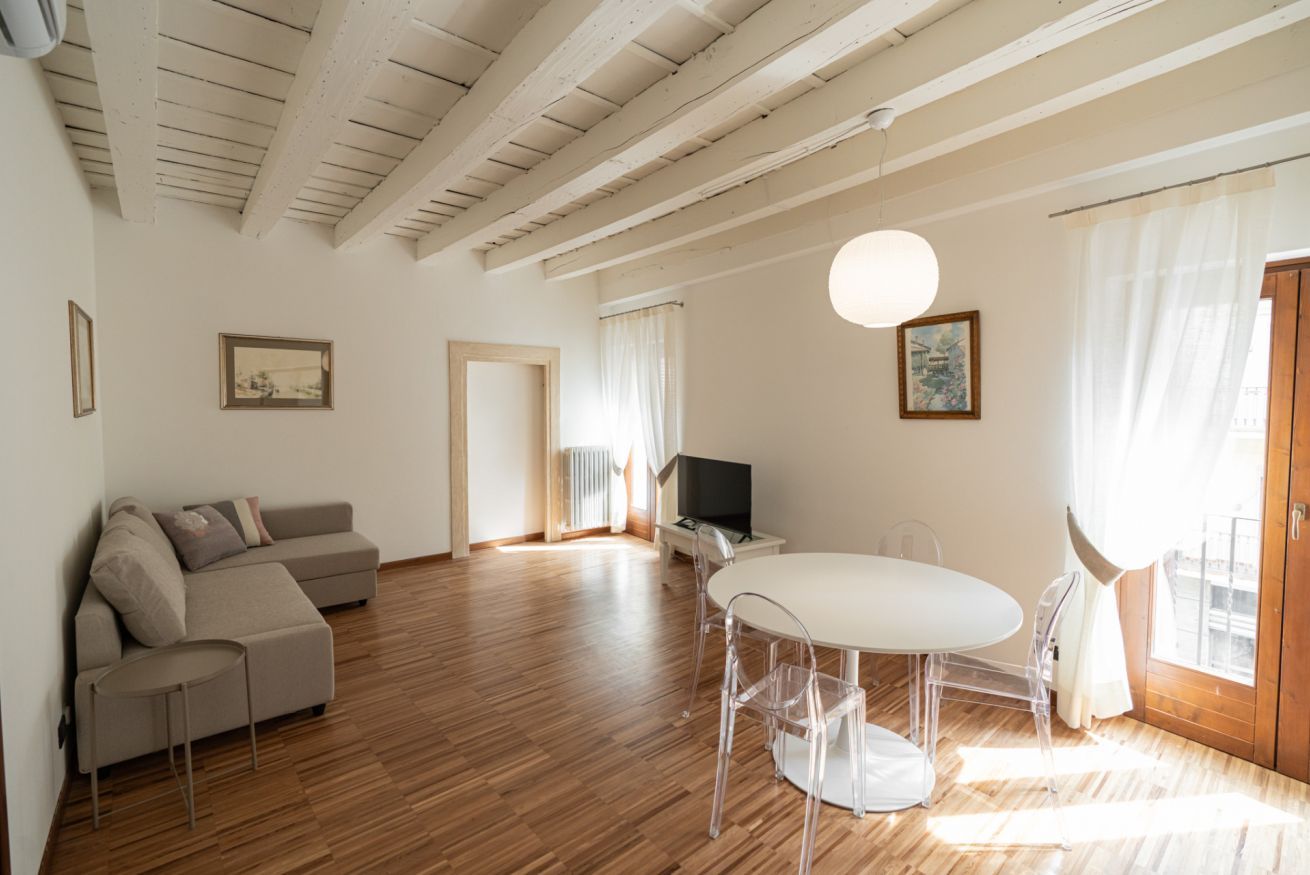A modern living room with a wooden floor, a beige sofa, a round dining table with clear chairs, a TV, and a large window with curtains.
