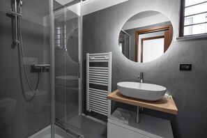 Un baño moderno con cabina de ducha de vidrio, un lavabo blanco sobre encimera de madera y un espejo redondo en la pared.